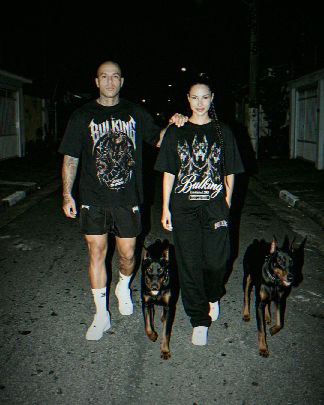Dupla com camisetas DARK ATDV e dois dobermans na rua, à noite